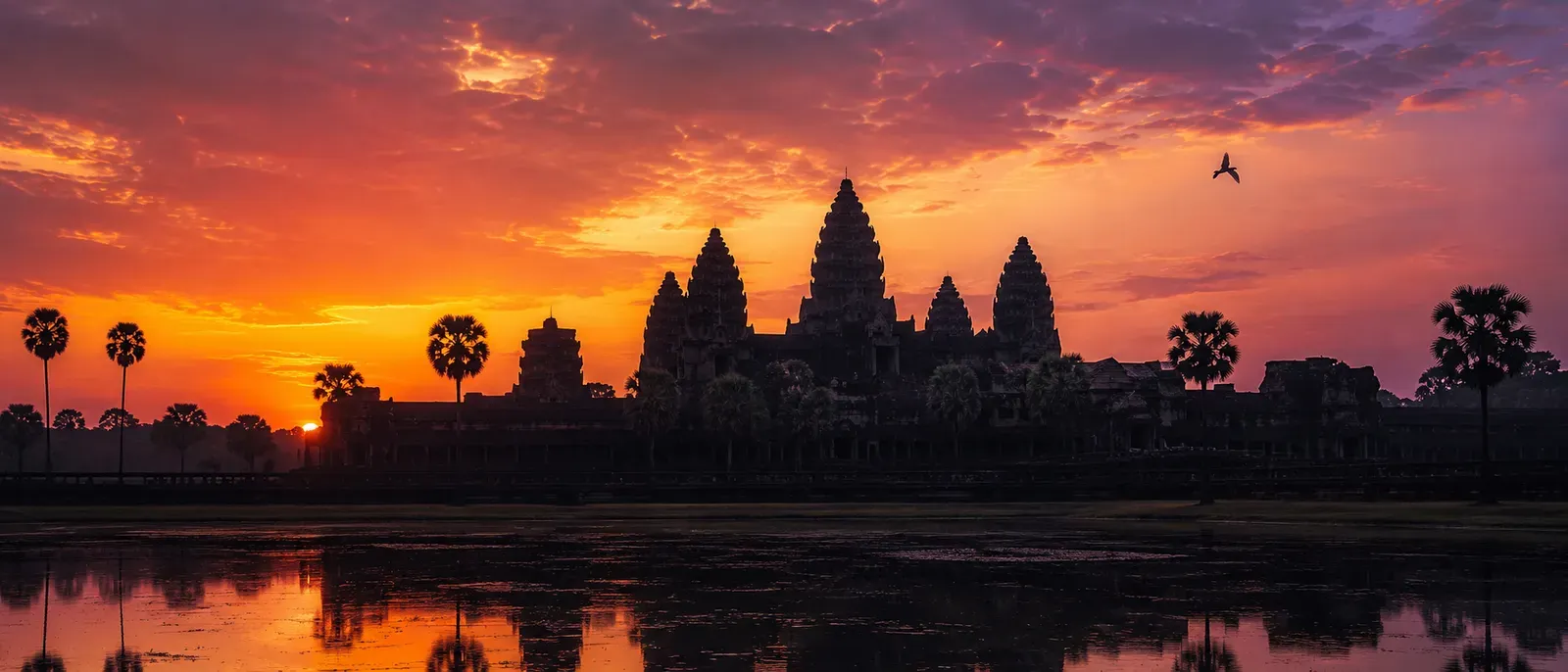 The spires of Angkor Wat in silhouette against a warm sunrise sky, mirrored in the water before it — a Vaiṣṇava temple in Cambodia.